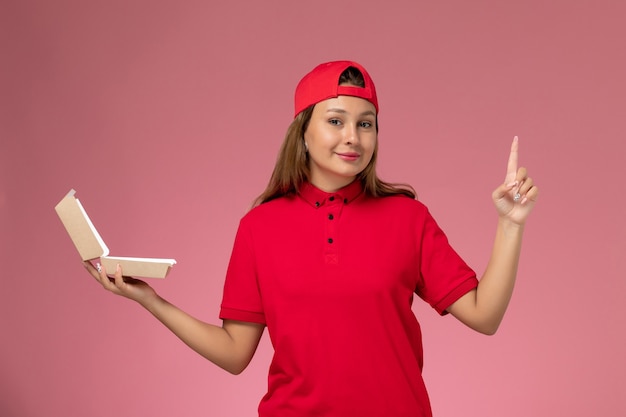 Weiblicher Kurier der Vorderansicht in der roten Uniform und im Umhang, die leeres Liefernahrungsmittelpaket auf hellrosa Wand halten, einheitliche Lieferservice-Firmenarbeit