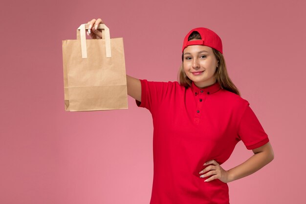 Weiblicher Kurier der Vorderansicht in der roten Uniform und im Umhang, die Lieferpapierpakete auf rosa Wand halten, Jobuniform-Lieferservice
