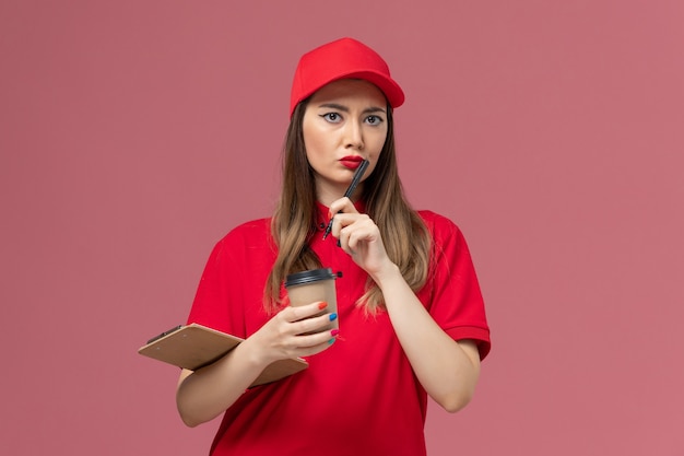 Weiblicher Kurier der Vorderansicht in der roten Uniform und im Umhang, die Lieferung Kaffeetasse mit Notizblock und Stift auf der hellrosa Hintergrunddienst-Joblieferuniform halten
