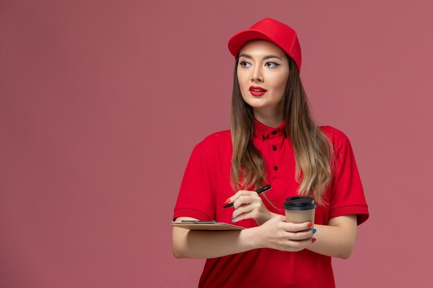 Weiblicher Kurier der Vorderansicht in der roten Uniform und im Umhang, die Lieferung Kaffeetasse mit Notizblock und Stift auf der hellrosa Hintergrunddienst-Joblieferuniform halten