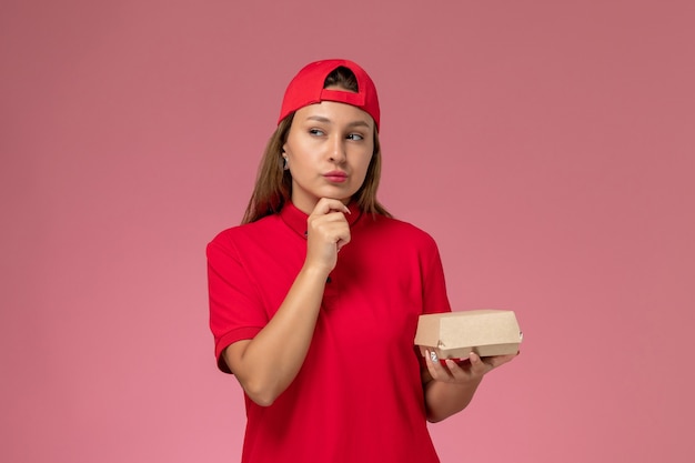 Weiblicher Kurier der Vorderansicht in der roten Uniform und im Umhang, die Lieferung-Nahrungsmittelpaket denken, das an der rosa Wand, Uniform-Lieferservice-Firmenjob denkt