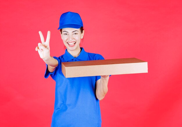 Weiblicher Kurier in blauer Uniform, der einen Pizzakarton zum Mitnehmen aus Pappe hält und ein Genusszeichen zeigt.