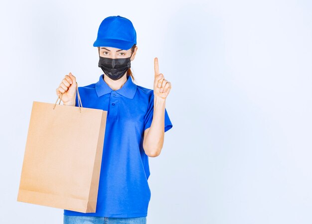 Weiblicher Kurier in blauer Uniform und Gesichtsmaske, der eine Einkaufstüte aus Karton hält und eine Idee oder ein Denken hat.