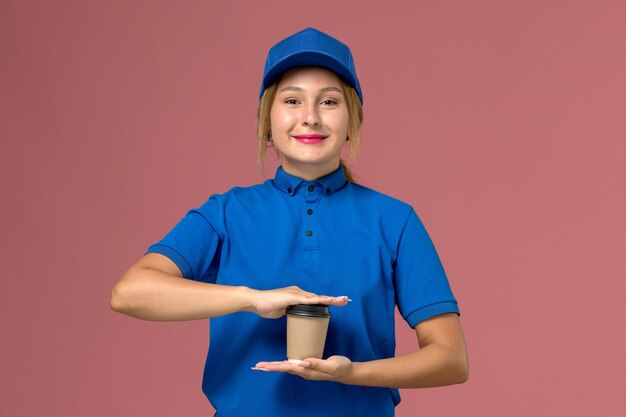 weiblicher Kurier in der blauen Uniform, die die Tasse Kaffee mit leichtem Lächeln auf rosa, Dienstuniform-Lieferjobarbeiter hält