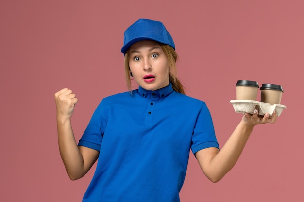 weiblicher Kurier in der blauen Uniform, die Lieferbecher des Kaffees hält, der auf rosa, Dienstarbeiterjobuniformlieferung aufwirft