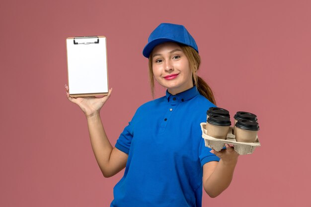 weiblicher Kurier in der blauen Uniform, die Notizblock zusammen mit braunen Lieferbechern des Kaffees hält, der auf hellrosa, Arbeitsdienstuniformlieferung lächelt