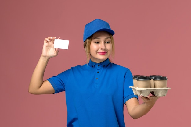 weiblicher Kurier in der blauen Uniform, die weiße Karte zusammen mit braunen Lieferbechern Kaffee auf hellrosa, Dienstjobuniformlieferung hält