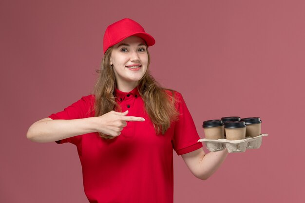 weiblicher Kurier in der roten Uniform, die braune Kaffeetassen der Lieferung hält und auf rosa, einheitlicher Arbeiterservicezustellung lächelt