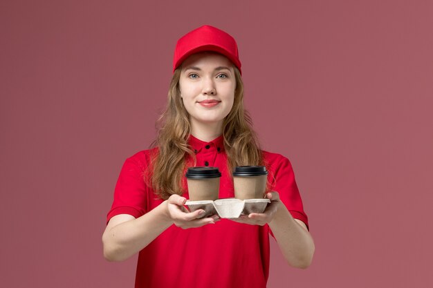 weiblicher Kurier in der roten Uniform, die die Kaffeetassen der Lieferung hält, die auf hellrosa, Dienstuniform-Servicearbeiterlieferung lächeln