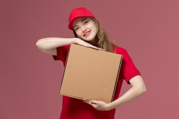 weiblicher Kurier in der roten Uniform, die Nahrungsmittellieferbox hält lächelnd auf rosa, einheitlicher Dienstlieferungsjob