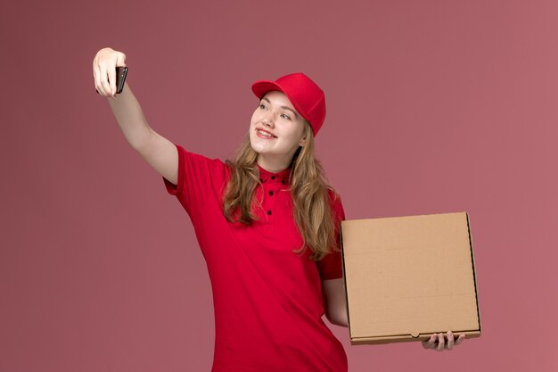 weiblicher Kurier in der roten Uniform, die Selfie mit Nahrungsmittelbox auf rosa, einheitlichem Arbeitsdienstarbeiter hält