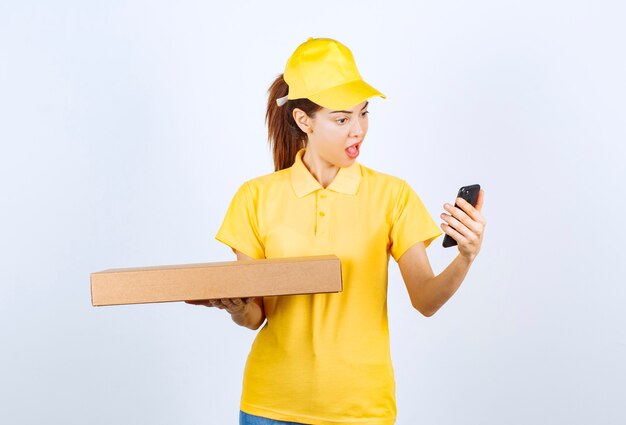 Weiblicher Kurier in gelber Uniform, der ein Papppaket hält, während er ihr Telefon überprüft und lächelt.