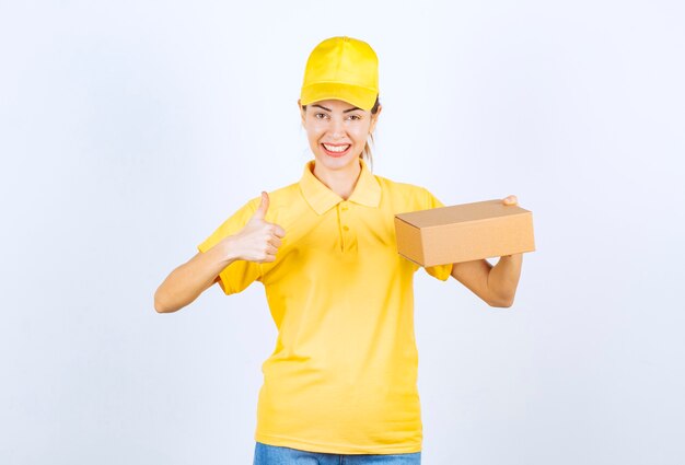 Weiblicher Kurier in gelber Uniform, der ein Papppaket liefert und ein erfolgreiches Handzeichen zeigt.