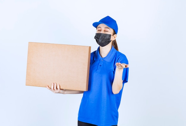 Weiblicher Kurier in Maske und blauer Uniform, der ein großes Papppaket hält und das Produkt riecht