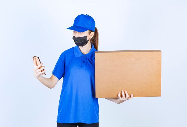 Weiblicher Kurier in Maske und blauer Uniform, der ein großes Papppaket hält und neue Bestellungen an ihrem Telefon entgegennimmt