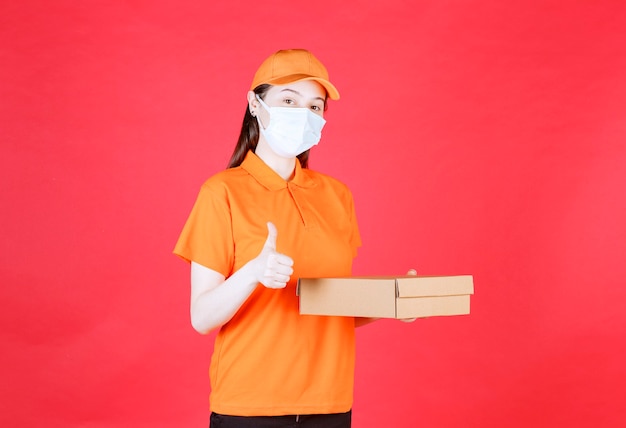 Weiblicher Kurier in orange Farbe Uniform und Maske hält einen Karton und zeigt positives Handzeichen.