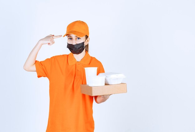 Weiblicher Kurier in orangefarbener Uniform und schwarzer Maske mit einem Papppaket, Essen und Getränken zum Mitnehmen beim Denken und Planen