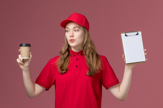 weiblicher Kurier in roter Uniform, die braune Lieferkaffeetasse und Notizblock auf rosa, einheitlichem Dienstlieferungsarbeiter hält