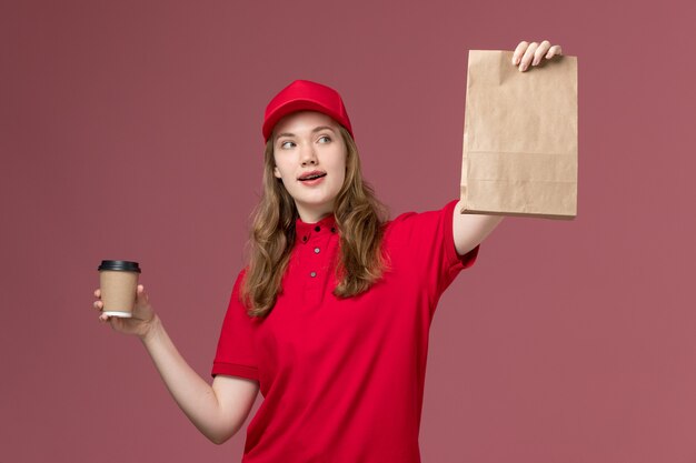 weiblicher Kurier in roter Uniform, die Lieferkaffeetasse und Lebensmittelpaket auf hellrosa, einheitliches Jobdienstarbeiter-Liefermädchen hält