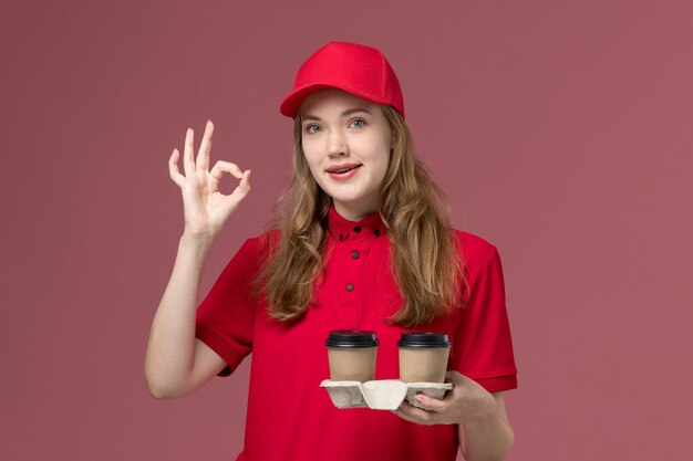 weiblicher Kurier in roter Uniform, die Lieferkaffeetassen auf hellrosa, einheitliches Jobdienstarbeiter-Liefermädchen hält
