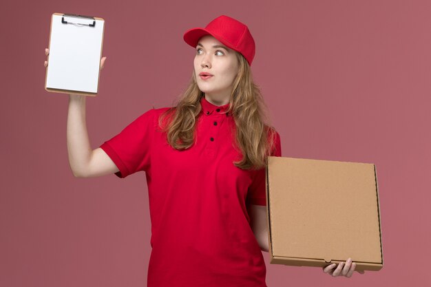 weiblicher Kurier in roter Uniform, die Nahrungsmittelbox mit Notizblock auf rosa, einheitlichem Dienstlieferungsarbeiter hält