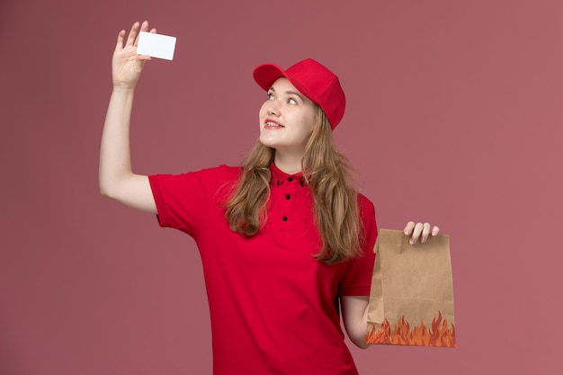 weiblicher Kurier in roter Uniform, die weiße Karte und Lebensmittelpaket auf hellrosa, Arbeitsuniform-Servicearbeiterlieferung hält