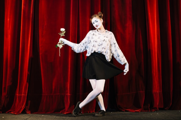 Weiblicher Pantomimekünstler, der mit dem gekreuzten Bein hält Weißrose steht