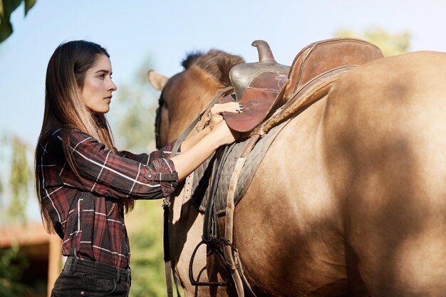 Weiblicher Pferdetrainer, der den Sattel vorbereitet, um an einem sonnigen Herbsttag auf einer Ranch zu reiten und von Prärien zu träumen