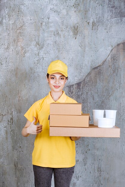 Weiblicher Servicemitarbeiter in gelber Uniform, die einen Vorrat an Pappkartons zum Mitnehmen und Plastikbecher hält.