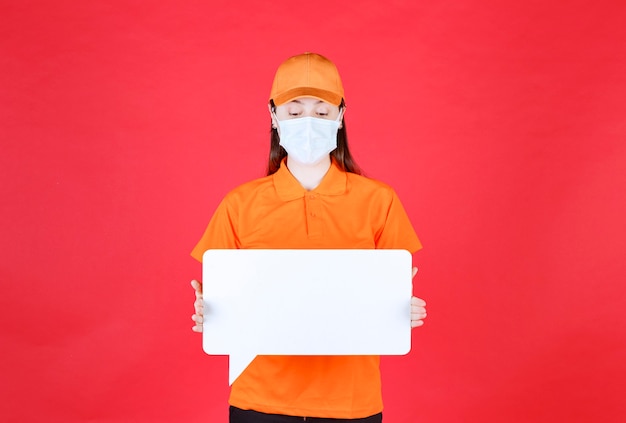 Weiblicher Servicemitarbeiter in orangefarbener Uniform und Maske, die eine weiße rechteckige Infotafel halten.