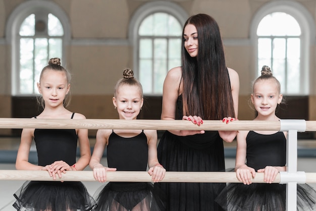 Weiblicher Trainer, der drei Ballerinamädchen stehen hinter dem Barre betrachtet