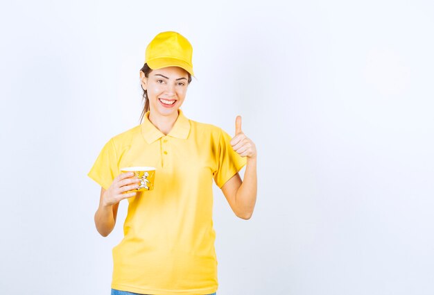 Weibliches Mädchen in gelber Uniform, das eine gelbe Nudelschale zum Mitnehmen hält und Freudenhandzeichen zeigt.