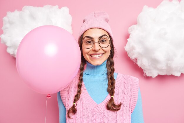 weibliches Model lächelt glücklich in Freizeitkleidung gekleidet hat zwei gekämmte lange Zöpfe trägt runde Brille hält aufgeblasenen Ballon vorweg für den Urlaub oder wenn die Party beginnt