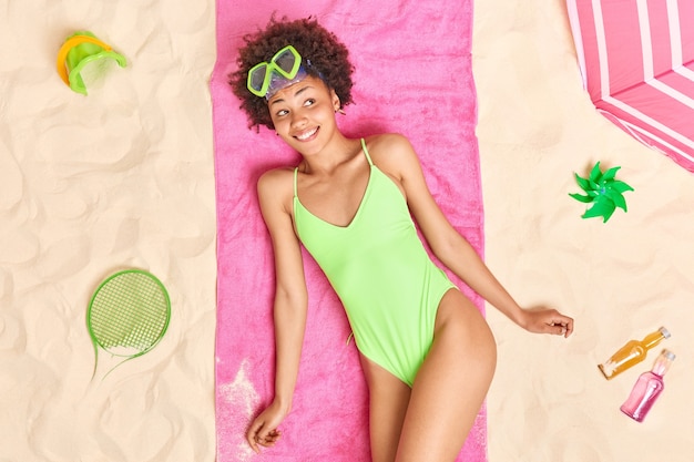 weibliches modell in grünem bikini trägt schnorchelmaske subathes während des sonnigen tages pses am sandstrand, umgeben von verschiedenen gegenständen. Sommerzeit und Ruhekonzept