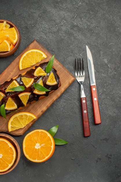 Weiche Kuchen auf Holzschneidebrett und Orangen mit Blattkeksen auf dunklem Tisch schneiden