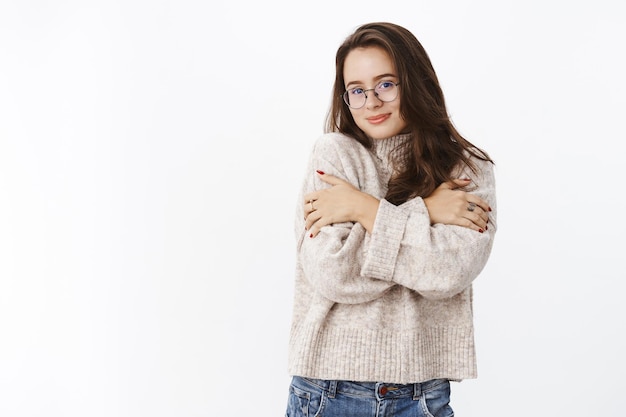 Weiche und zarte glückliche junge sinnliche Frau in Brille und Pullover, die sich zum Aufwärmen in gemütlichem Outfit umarmt