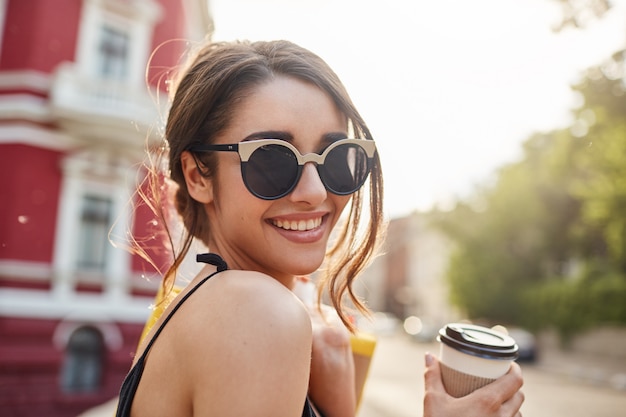 Weicher Fokus. Lifestyle-Konzept. Schließen Sie herauf Porträt der freudigen jungen attraktiven dunkelhaarigen kaukasischen Frau in der Sonnenbrille und im schwarzen Outfit, die mit Zähnen lächeln, Kaffee trinken, Taschen mit Kleidern af halten