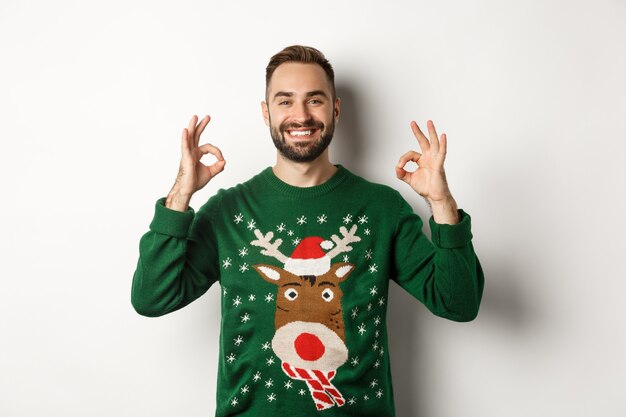 Weihnachten, Feiertage und Feiern. Zufriedener lächelnder Mann im grünen Pullover, der OK-Zeichen zeigt und zustimmend nickt, Produkt empfehlend, auf weißem Hintergrund stehend.