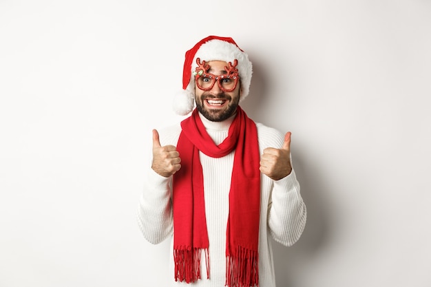 Weihnachten, Neujahr und Feierkonzept. Aufgeregter Mann mit Weihnachtsmütze und Partybrille, Daumen hoch zur Zustimmung, zufrieden lächelnd, weißer Hintergrund