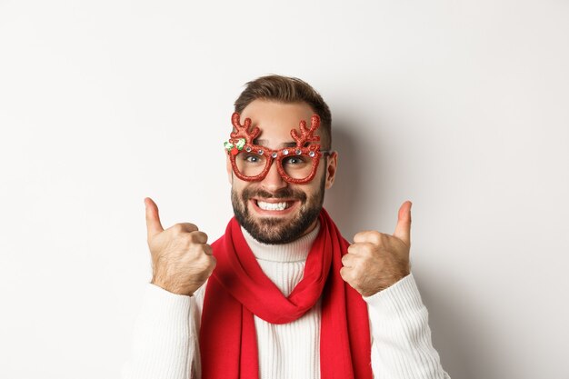 Weihnachten, Neujahr und Feierkonzept. Glücklicher und zufriedener Mann mit Bart, der eine Partybrille trägt, Daumen hoch zeigt oder wie, auf weißem Hintergrund stehend