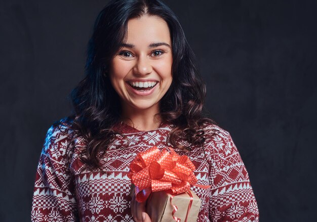 Weihnachten, Valentinstag, Neujahr. Porträt eines glücklichen brünetten Mädchens, das einen warmen Pullover trägt und eine Geschenkbox hält, isoliert auf einem dunklen, strukturierten Hintergrund.
