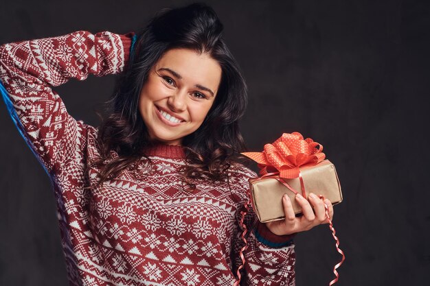 Weihnachten, Valentinstag, Neujahr. Porträt eines glücklichen brünetten Mädchens, das einen warmen Pullover trägt und eine Geschenkbox hält, isoliert auf einem dunklen, strukturierten Hintergrund.