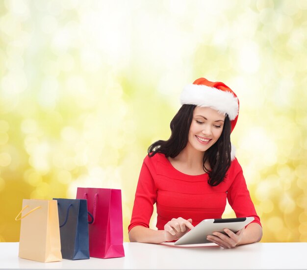 Weihnachten, weihnachten, online-shopping und elektronikkonzept - lächelnde frau in nikolausmütze mit einkaufstüten und tablet-pc