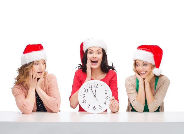 Weihnachten, weihnachten, winter, glückskonzept - drei lächelnde frauen in weihnachtsmannmützen mit uhr mit 12
