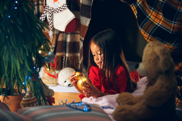 Weihnachts-, Feiertags- und Kindheitskonzept - glückliches Mädchen in den roten Kleidern, die natürlichen Tannenbaum verzieren