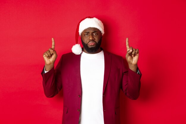 Weihnachts-, Party- und Feiertagskonzept. Elender und trauriger Afroamerikaner, der mit den Fingern nach oben zeigt, enttäuscht aussieht, Weihnachtsmütze trägt, roter Hintergrund.
