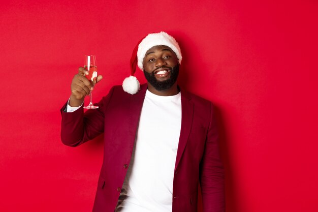 Weihnachts-, Party- und Feiertagskonzept. Fröhlicher schwarzer Mann, der Prost sagt, ein Glas Champagner anhebt und ein frohes neues Jahr wünscht, vor rotem Hintergrund stehend.