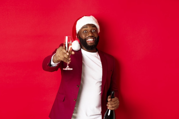 Weihnachts-, Party- und Feiertagskonzept. Schöner schwarzer Mann in Weihnachtsmütze, der ein Glas Champagner anhebt und lächelt, Toast sagt, Neujahr feiert, roter Hintergrund.