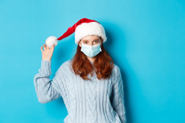 Weihnachts-, Quarantäne- und Covid-19-Konzept. Rothaariges Mädchen, das Gesichtsmaske trägt und mit Weihnachtsmütze spielt, Neujahr bei Lockdown feiert und auf blauem Hintergrund steht.