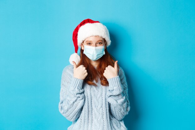 Weihnachts-, Quarantäne- und Covid-19-Konzept. Süßes rothaariges Teenie-Mädchen in Weihnachtsmütze und Pullover, Gesichtsmaske aus Coronavirus tragend, Daumen nach oben zeigend, auf blauem Hintergrund stehend